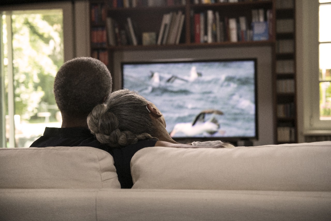 A couple wearing BTE Hearing Aids is watching TV on the sofa